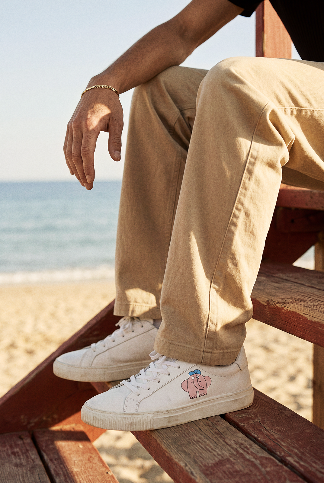 Close crop of white sneakers with elephant artwork, beige trousers and a gold bracelet on red wooden steps.