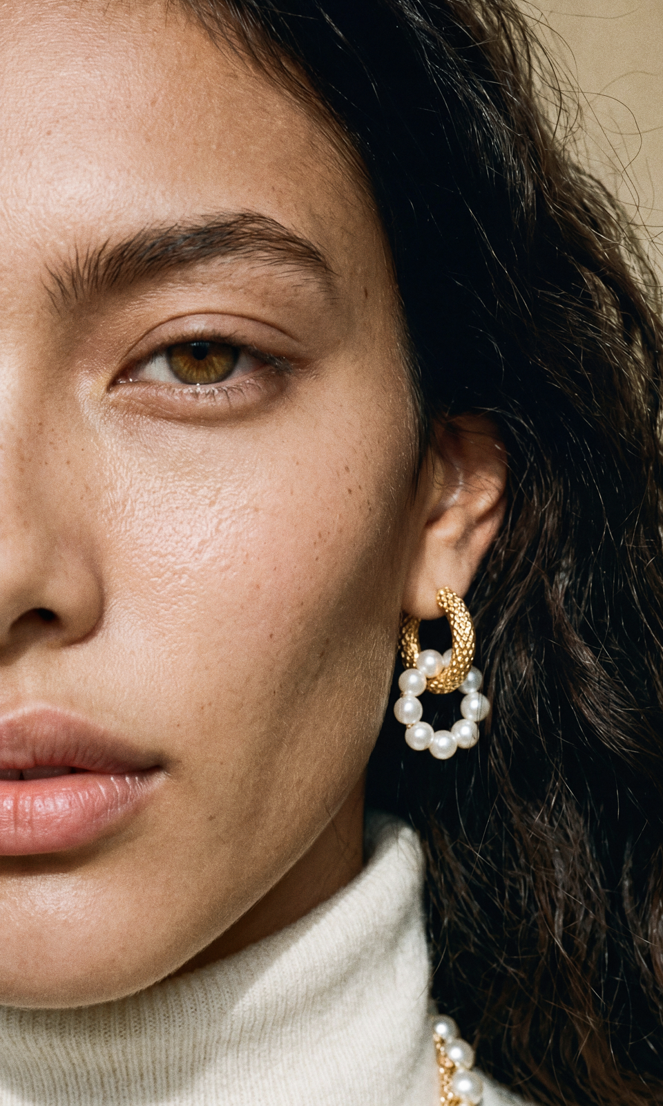 Tight macro crop of a gold-and-pearl hoop earring against soft skin and dark hair, lit with warm sunlight.