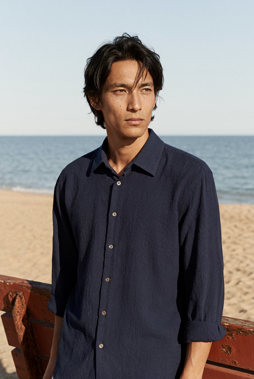 Editorial portrait of a man in a navy shirt standing by the water and weathered red structure.