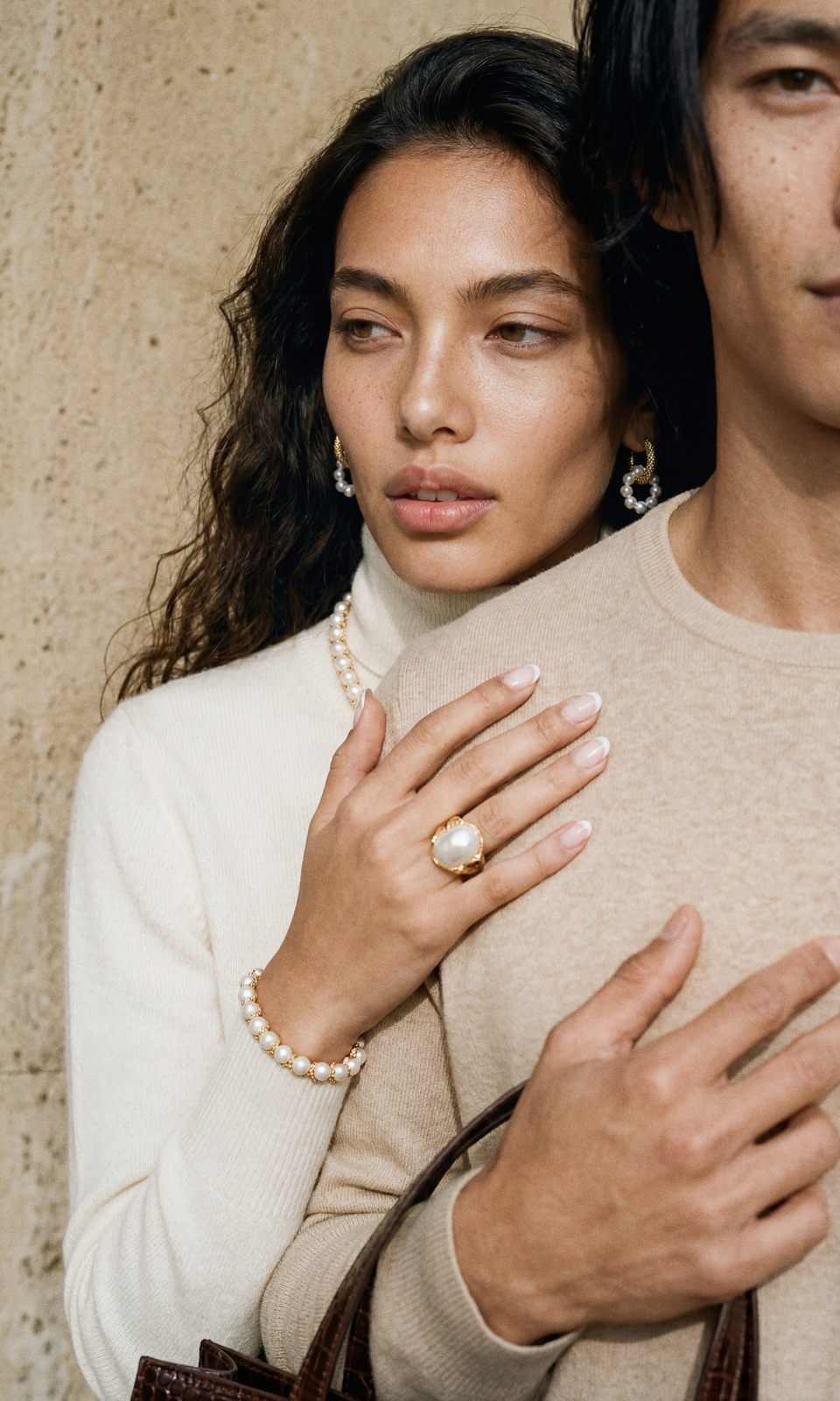 Two models in cream cashmere against weathered stone — pearl hoops, a baroque pearl ring and a pearl bracelet, with a brown croc tote in the foreground.