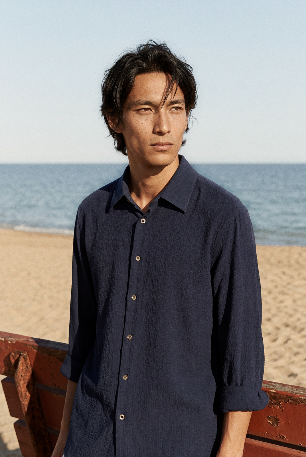 Editorial portrait of a man in a navy shirt standing by the water and weathered red structure.