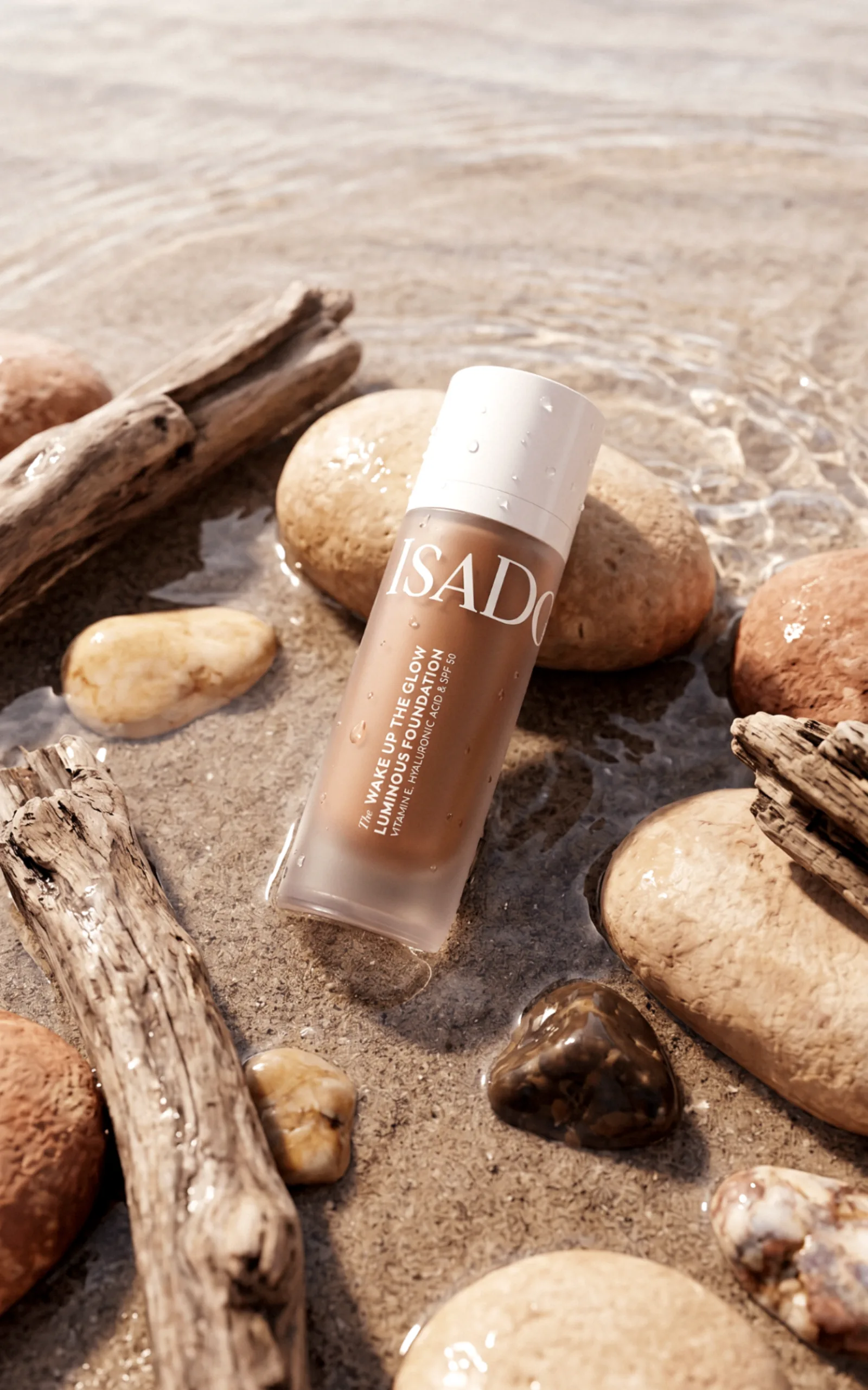 IsaDora Wake Up the Glow foundation — deeper shade lying on wet shore stones with water droplets.